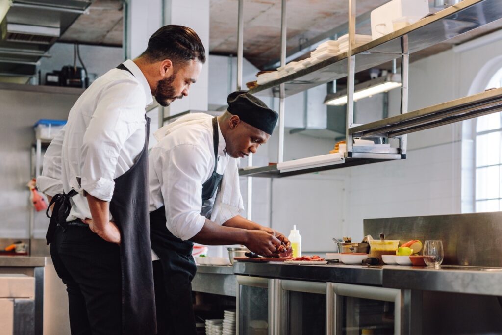 Chef meticulously preparing food in a commercial kitchen with a supervisor observing the process to showcase the importance of Protecting Your Restaurant with Food Contamination Insurance