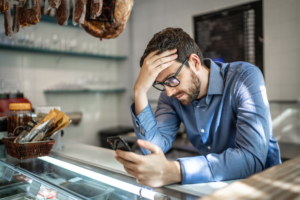 Stressed business owner in a deli, leaning on the counter while looking at his phone, reflecting challenges and pressures of managing a small business.